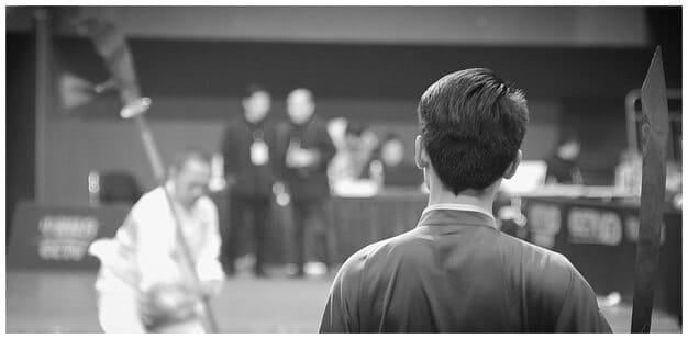 A behind-the-scenes photograph of a young Kang Ha Neul during a rehearsal for a school theater production. He is dressed in casual clothes, focused and engaged, with the director giving instructions in the background.